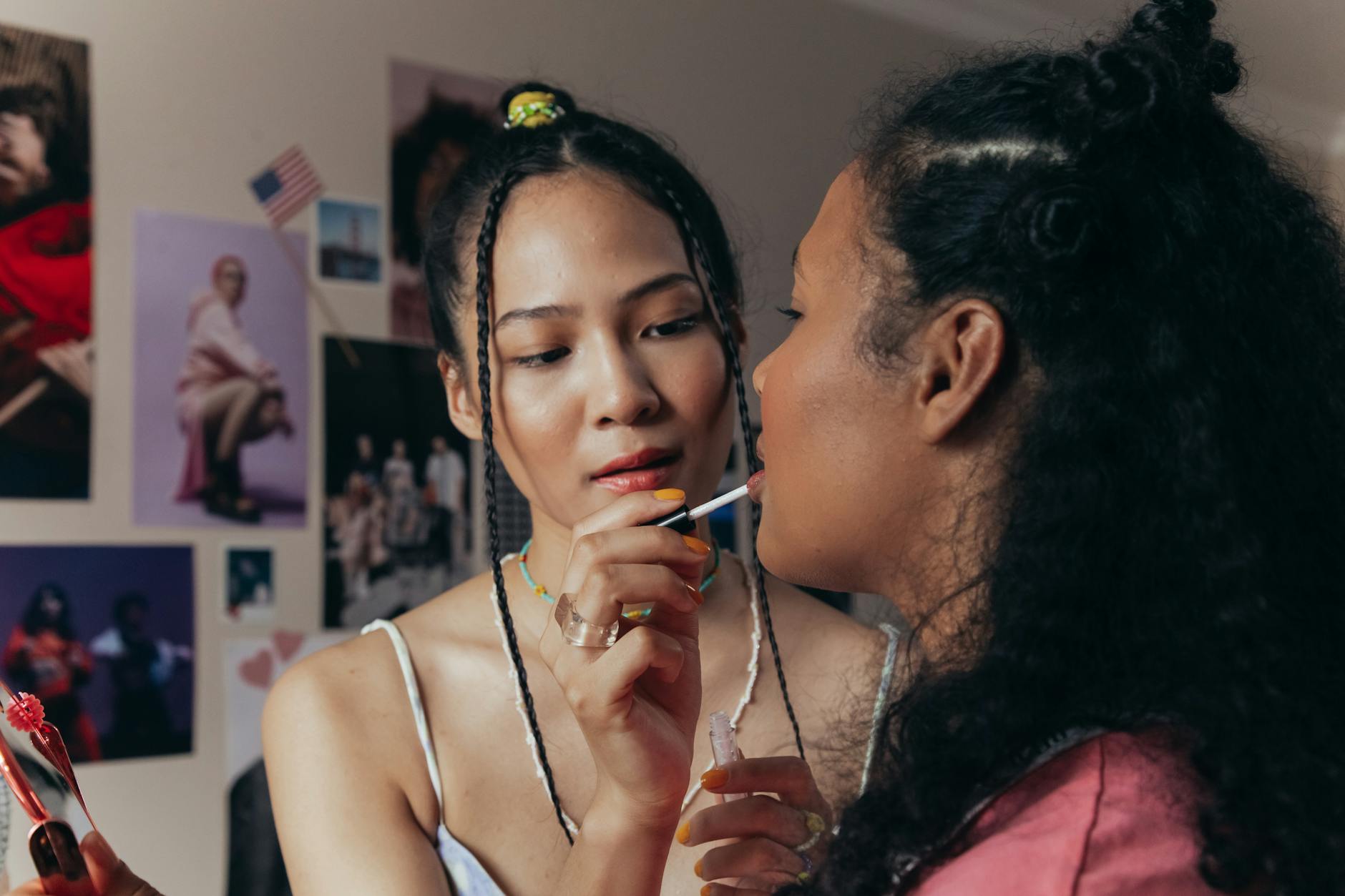 Two diverse women applying lipstick in a room adorned with posters, capturing a cozy and intimate moment.